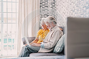 Pretty elderly 70s grey-haired couple resting on couch in living room hold on lap laptop watching movie smiling enjoy free time,