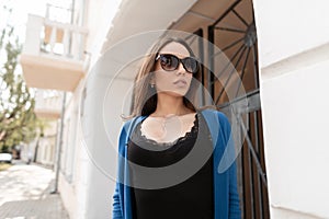 Pretty elegant modern young hipster woman in stylish sunglasses in a trendy top with lace in a blue knitted cape stands in a city