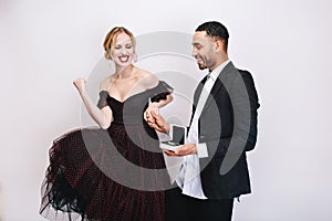 Pretty excited young woman in luxury dress expressing brightful happy emotions near handsome guy with gift on white