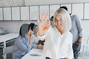 Pretty fair-haired female student posing with big smile and okay sign in front of group of international friends. Indoor