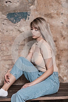 Pretty fashion young blonde woman in stylish blue jeans in a trendy shirt poses near a brick old wall indoors. Glamorous girl