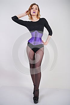 Pretty fashionable blonde girl wearing black jumpsuit with mesh pantyhose and purple corset on white background