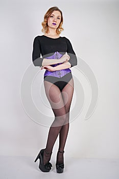 Pretty fashionable blonde girl wearing black jumpsuit with mesh pantyhose and purple corset on white background