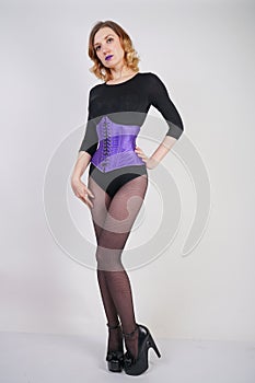 Pretty fashionable blonde girl wearing black jumpsuit with mesh pantyhose and purple corset on white background