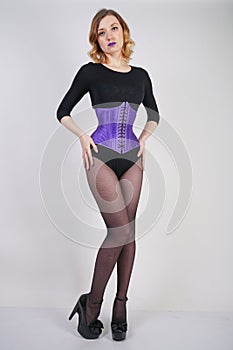 Pretty fashionable blonde girl wearing black jumpsuit with mesh pantyhose and purple corset on white background