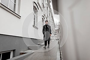 Pretty fashionable young man model in a gray elegant plaid coat with stylish hairstyle in autumn trendy boots walks near a vintage