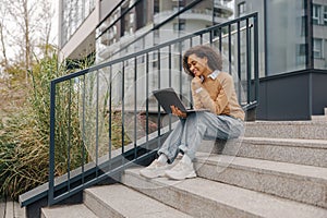 Pretty female entrepreneur working on laptop sitting outside on modern building background