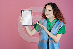 Pretty, female paramedic wearing vest, posing indoors, holding folder, showing, smiling.