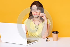 Pretty female sits at white desk with opened laptop computer, writes email, uses high speed internet, poses isolated over yellow