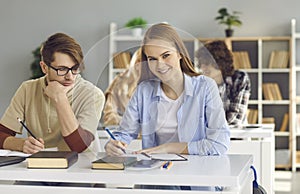 Pretty female student writing in notebook and looking at camera in classroom