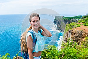 Pretty female traveler with a backpack standing on the edge of a cliff and smiling on camera. Bali, Indonesia