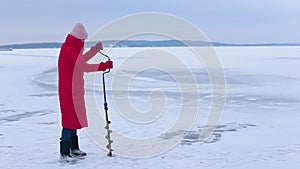 Pretty fisherwoman in red warm jacket and felt boots makes hole for ice fishing with help ice drill on frozen river