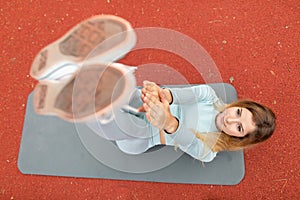 Pretty fit blond woman lying on yoga mat and reaching hands to her feet, training on the court outdoor on summer day