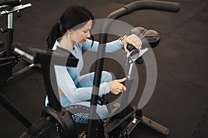 Pretty fitness woman adjusts the height of the exercise bike seat before starting a workout.