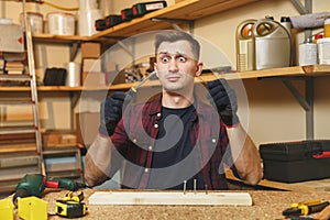 Handsome smiling young man working in carpentry workshop at wooden table place with piece of wood photo