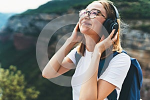 Pretty fun young tourist woman with backpack and  hipster glasses smiling listens favorite music with headphones enjoying freedom