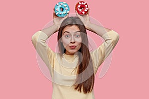 Pretty funny brunette young woman holds two delicious donuts, dressed in yellow clothes, models over pink background