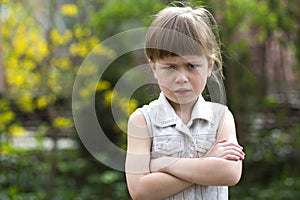 Pretty funny moody little blond preschool girl in white sleeveless dress looks into camera feeling angry and unsatisfied on blurre