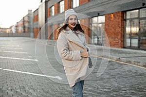 Pretty funny young woman in a fashionable knitted hat in a stylish coat of milk color from eco-fur stands and smiling near a brick