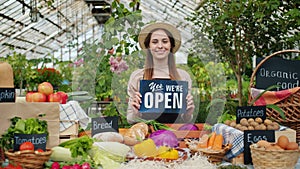Pretty girl in apron and hat holding open sign in organic food market smiling