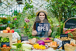 Pretty girl in apron and hat holding open sign in organic food market smiling