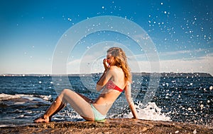 Pretty girl in a bikini beside the ocean laughing as she is splashed by a wave crashing on the rocks.