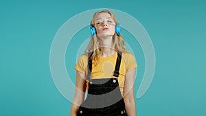 Pretty girl with blonde hair listening to music, smiling, dancing in headphones in studio against blue background. Music