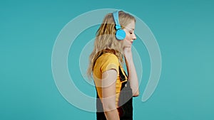 Pretty girl with blonde hair listening to music, smiling, dancing in headphones in studio against blue background. Music