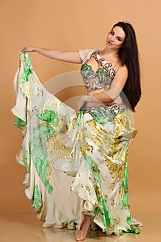 A pretty girl brunette who dances go-go and belly dance, stretching posing in studio in green arabian dress on a beige background