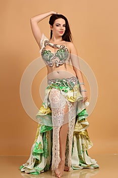 A pretty girl brunette who dances go-go and belly dance, stretching posing in studio in green arabian dress on a beige background