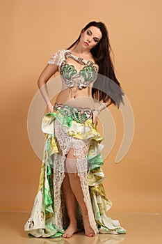 A pretty girl brunette who dances go-go and belly dance, stretching posing in studio in green arabian dress on a beige background