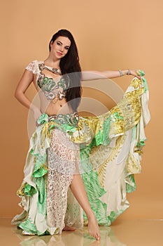 A pretty girl brunette who dances go-go and belly dance, stretching posing in studio in green arabian dress on a beige background