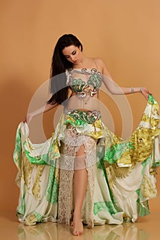 A pretty girl brunette who dances go-go and belly dance, stretching posing in studio in green arabian dress on a beige background