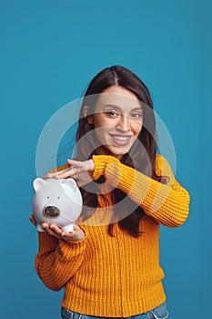 Pretty girl in casual orange sweater holding white piggy bank with lots of money