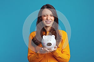 Pretty girl in casual orange sweater holding white piggy bank and winking