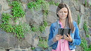 Pretty girl in a cotton jacket using tablet computer with earphones. Vintage wall of wild stone in the background.