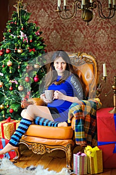 Pretty girl with a cup of coffee sits in a chair near the festive Christmas tree