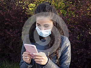 A pretty girl with curly hair in a medical mask is chatting on her phone in the green spring park