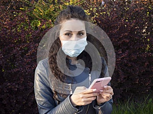 Pretty girl with curly hair in a medical mask with her smartphone in a green spring park