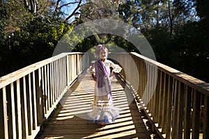 A pretty girl dancing flamenco in a typical gypsy dress with frills and fringes walks on a wooden bridge in a famous park in