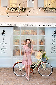 Pretty girl in a dress and with a straw hat is standing with a yellow retro bicycle in a European city. On the background of the c