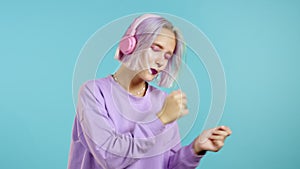 Pretty girl with dyed violet hair listening to music, smiling, dancing in headphones in studio against blue background