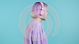 Pretty girl with dyed violet hair listening to music, smiling, dancing in headphones in studio against blue background