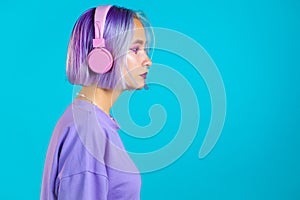 Pretty girl with dyed violet hair listening to music, smiling, dancing in headphones in studio against blue background