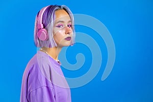 Pretty girl with dyed violet hair listening to music, smiling, dancing in headphones in studio against blue background