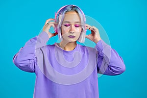 Pretty girl with dyed violet hair listening to music, smiling, dancing in headphones in studio against blue background