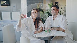 Pretty girl and her loving boyfriend are taking selfie with cocktail glasses using smartphone while relaxing in spa