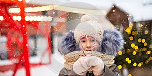 Pretty girl holds in his hands candy cane in the shape of a heart outdoor in warm clothes in winter festive market. Fairy lights