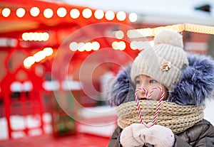 Pretty girl holds in his hands candy cane in the shape of a heart outdoor in warm clothes in winter festive market. Fairy lights
