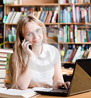 Pretty girl in library typing on laptop and talking on the phone